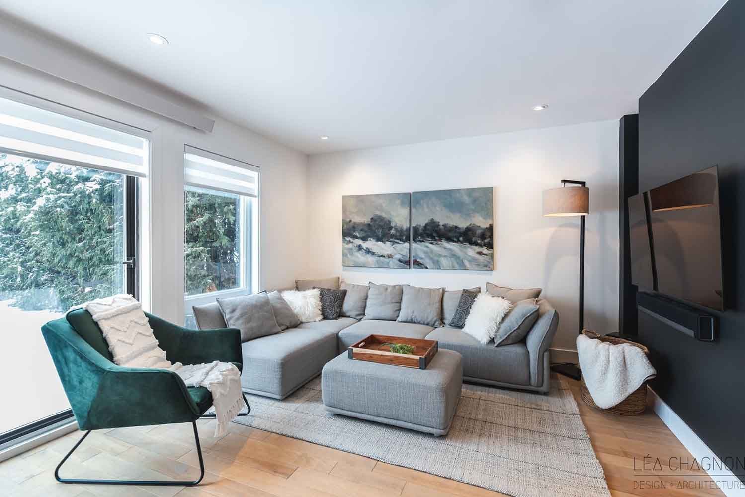 Salon moderne avec canapé d'angle gris, fauteuil vert et œuvre d'art au-dessus d'un pouf en bois.