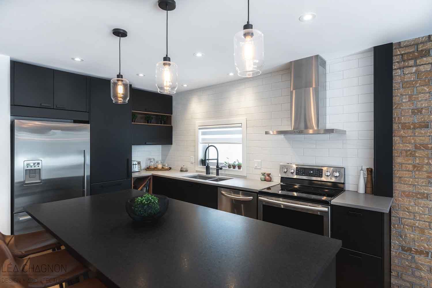 Cuisine moderne avec armoires noires, électroménagers en acier inoxydable, îlot central et luminaires suspendus.