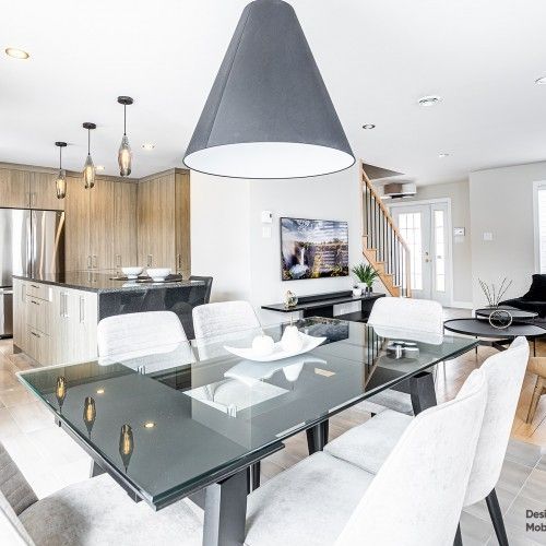 Salle à manger moderne avec table en verre, chaises blanches et grande suspension lumineuse grise.