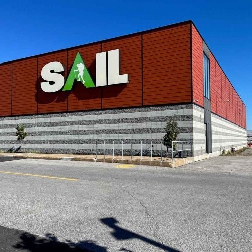 Extérieur d'un magasin SAIL avec un grand logo vert et blanc sur un bâtiment rouge et gris.