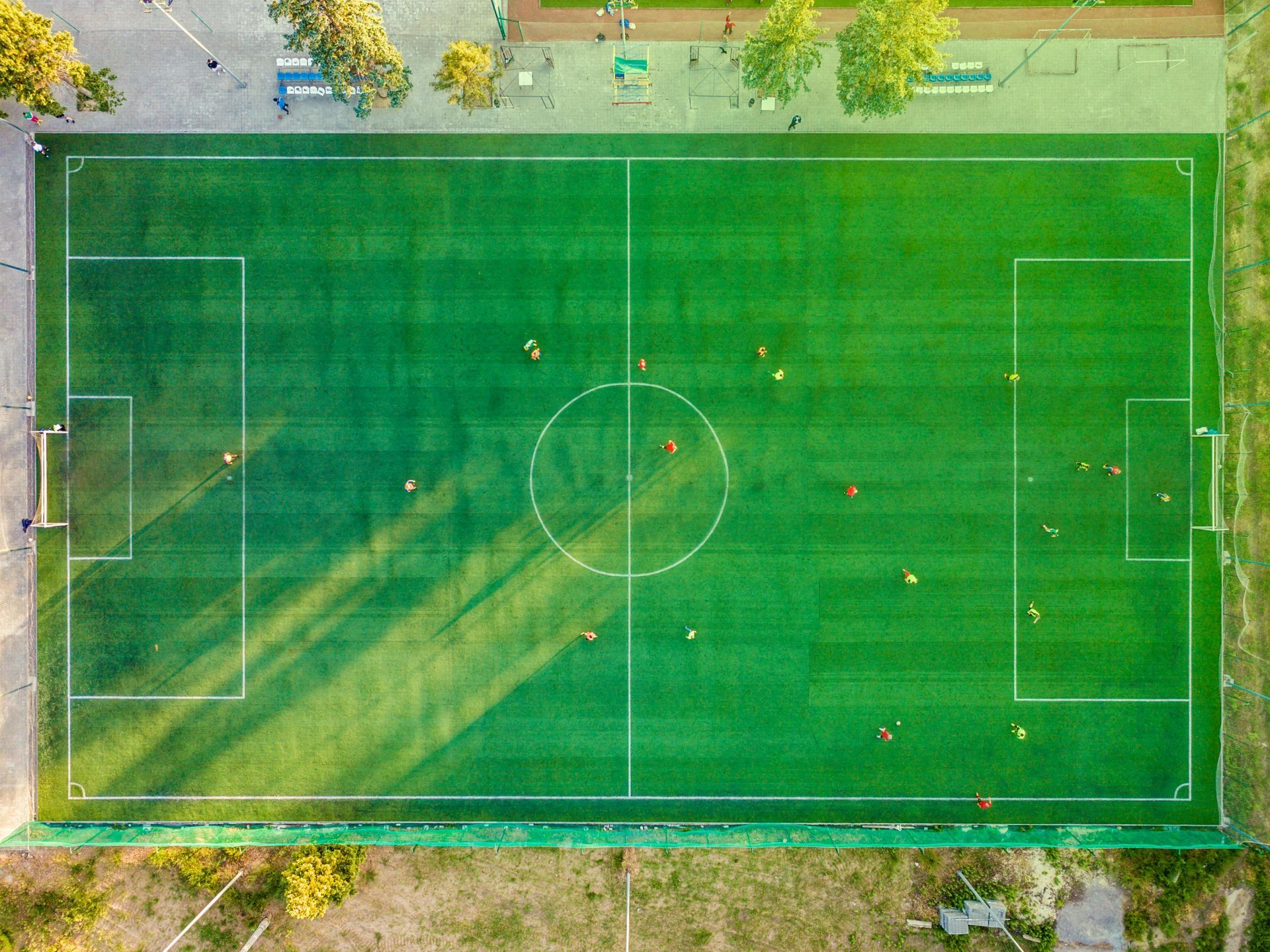Aerial view of a soccer field with players. Green grass, white lines, goal posts. Sunlight casts long shadows.