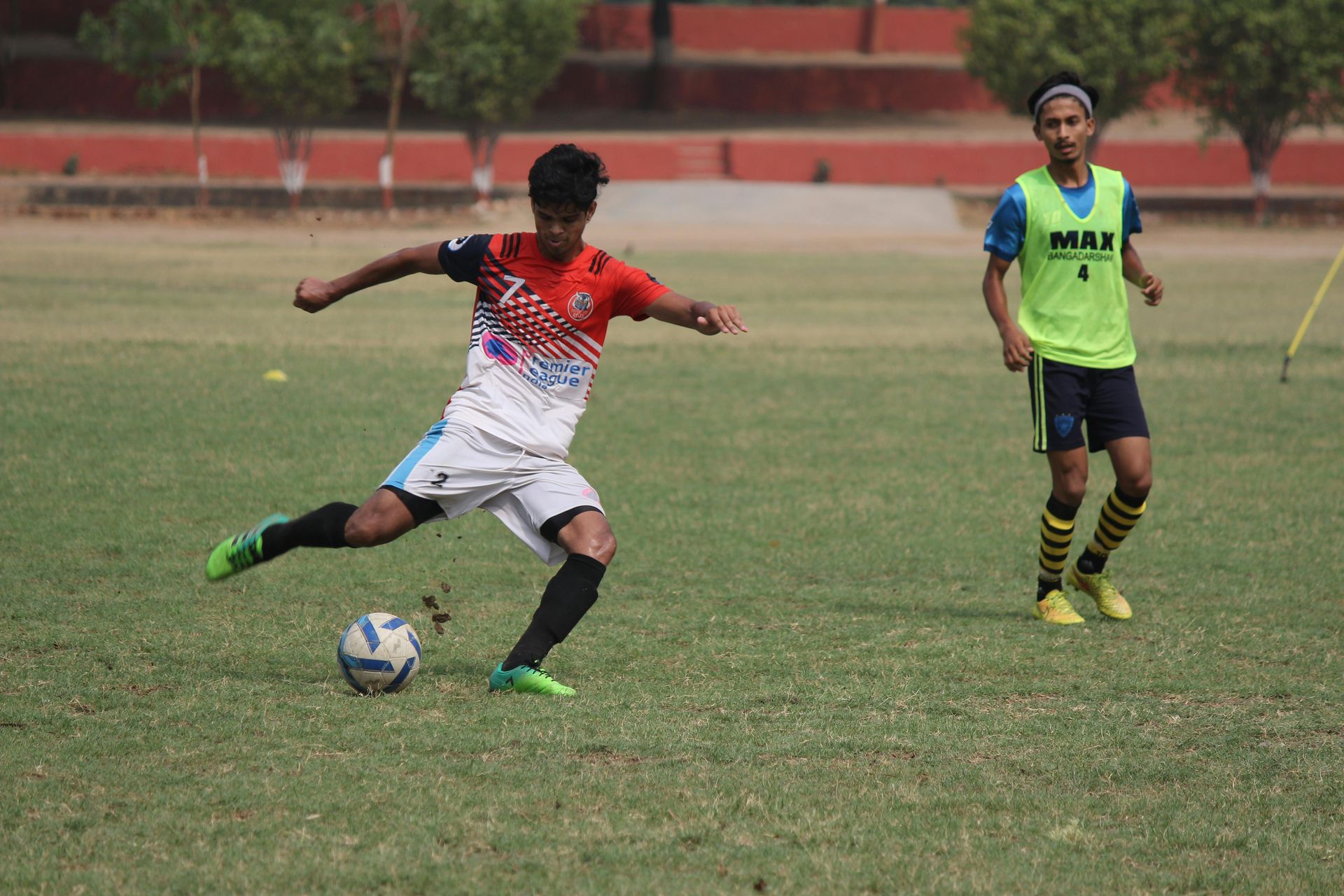 Soccer player in white, red and blue jersey kicks a ball on a green field. Another player watches in the background.