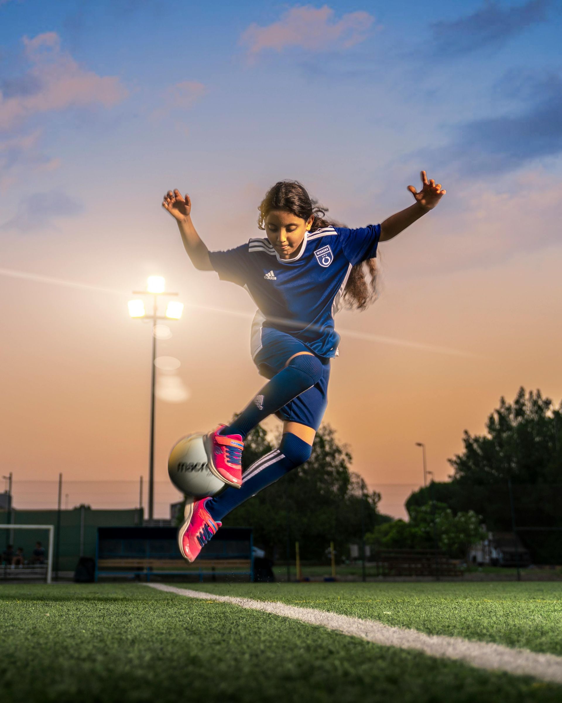Soccer player mid-air, kicking ball. Blue uniform, stadium setting, sunset sky.