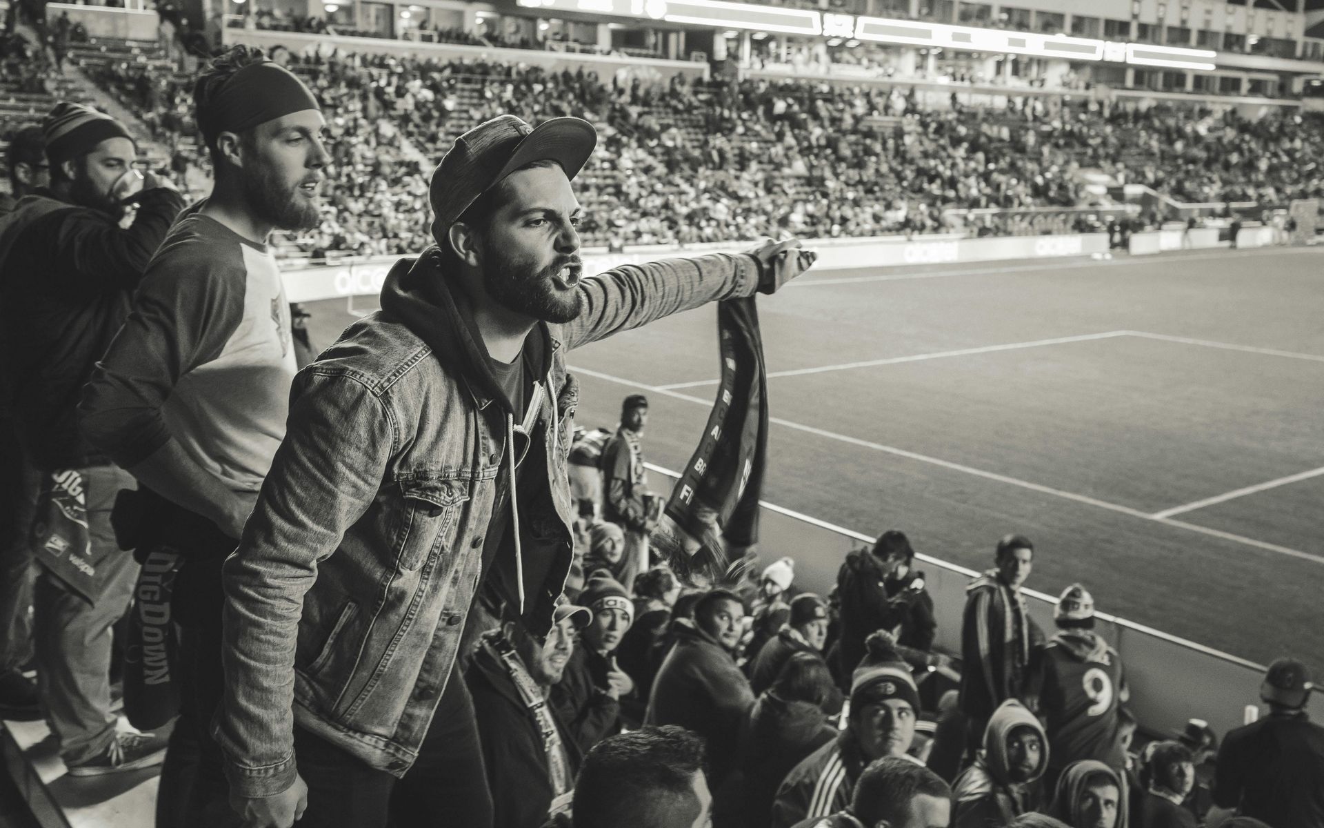 Man in a stadium cheering, holding a flag. Other fans are around him in the bleachers.