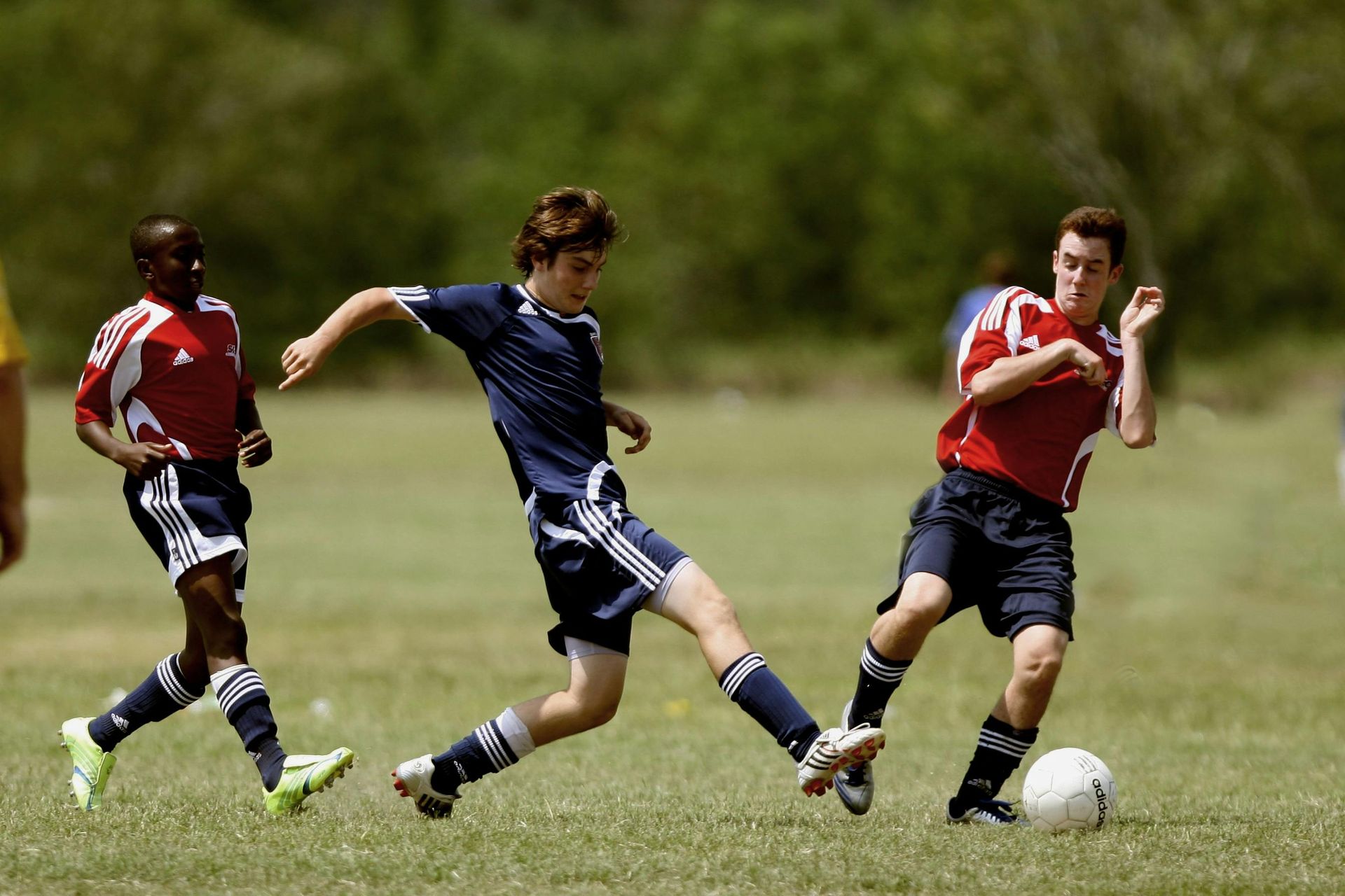 Soccer players in action on a grassy field. One player kicks the ball while two others run nearby.