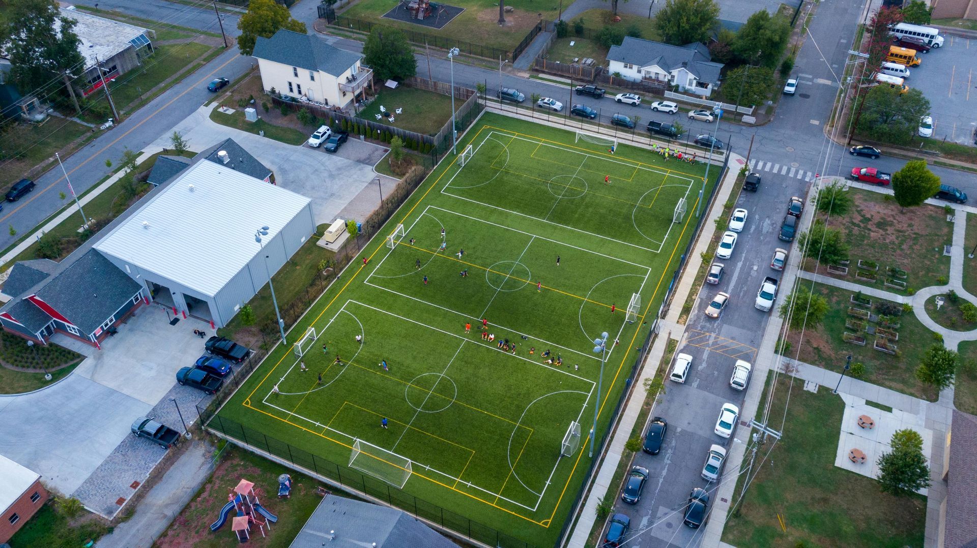 Aerial view of a green soccer field in a residential neighborhood, with surrounding streets and buildings.