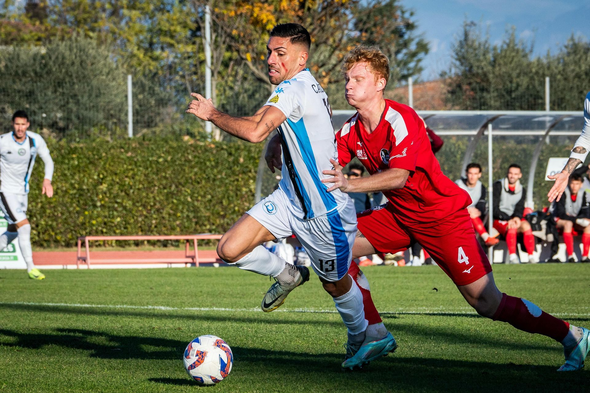 Soccer players, one in white and blue, the other in red, on a sunny field.