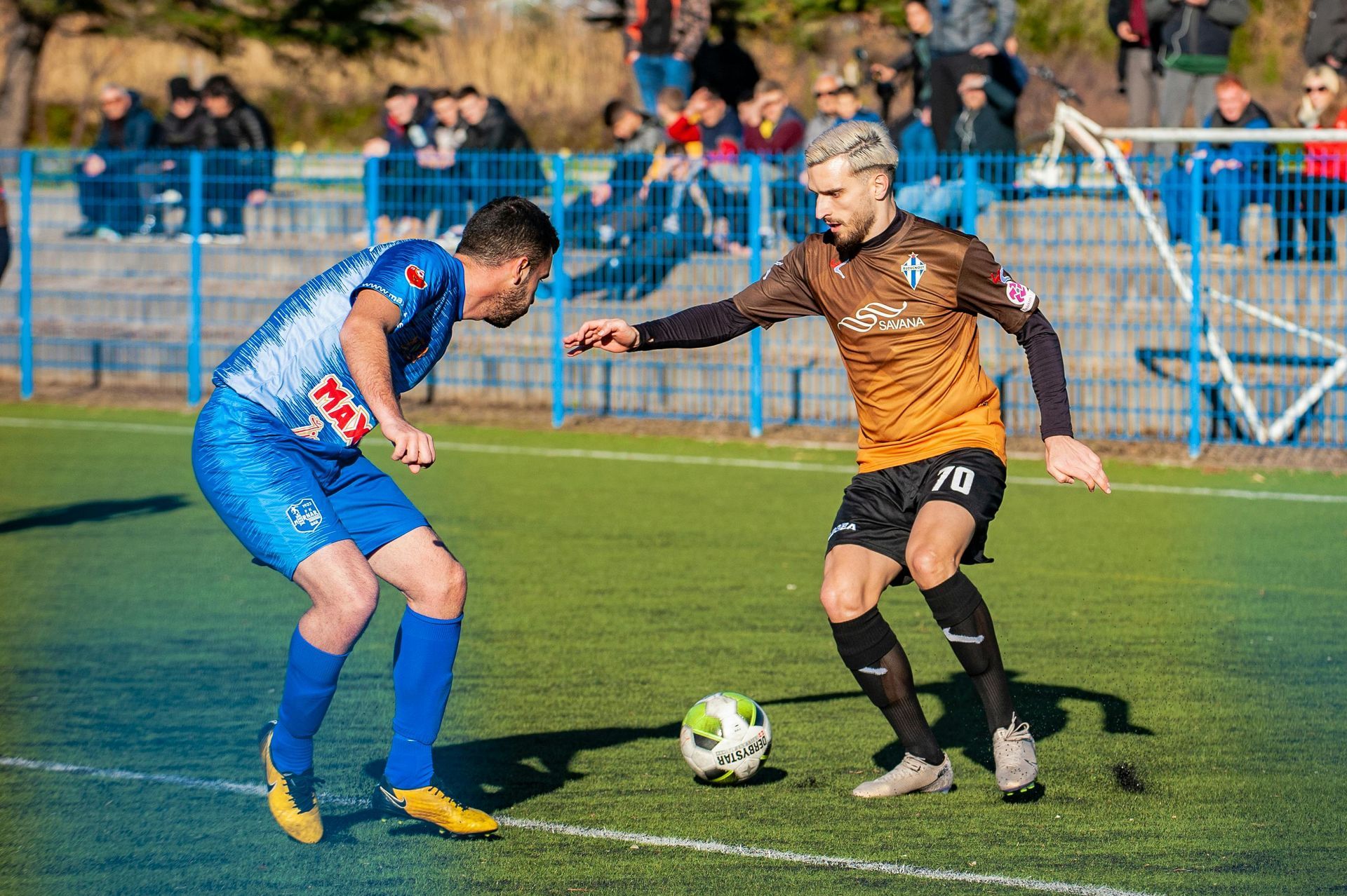 Soccer players in blue and orange uniforms on a green field, one dribbling, other defending. Spectators in the background.