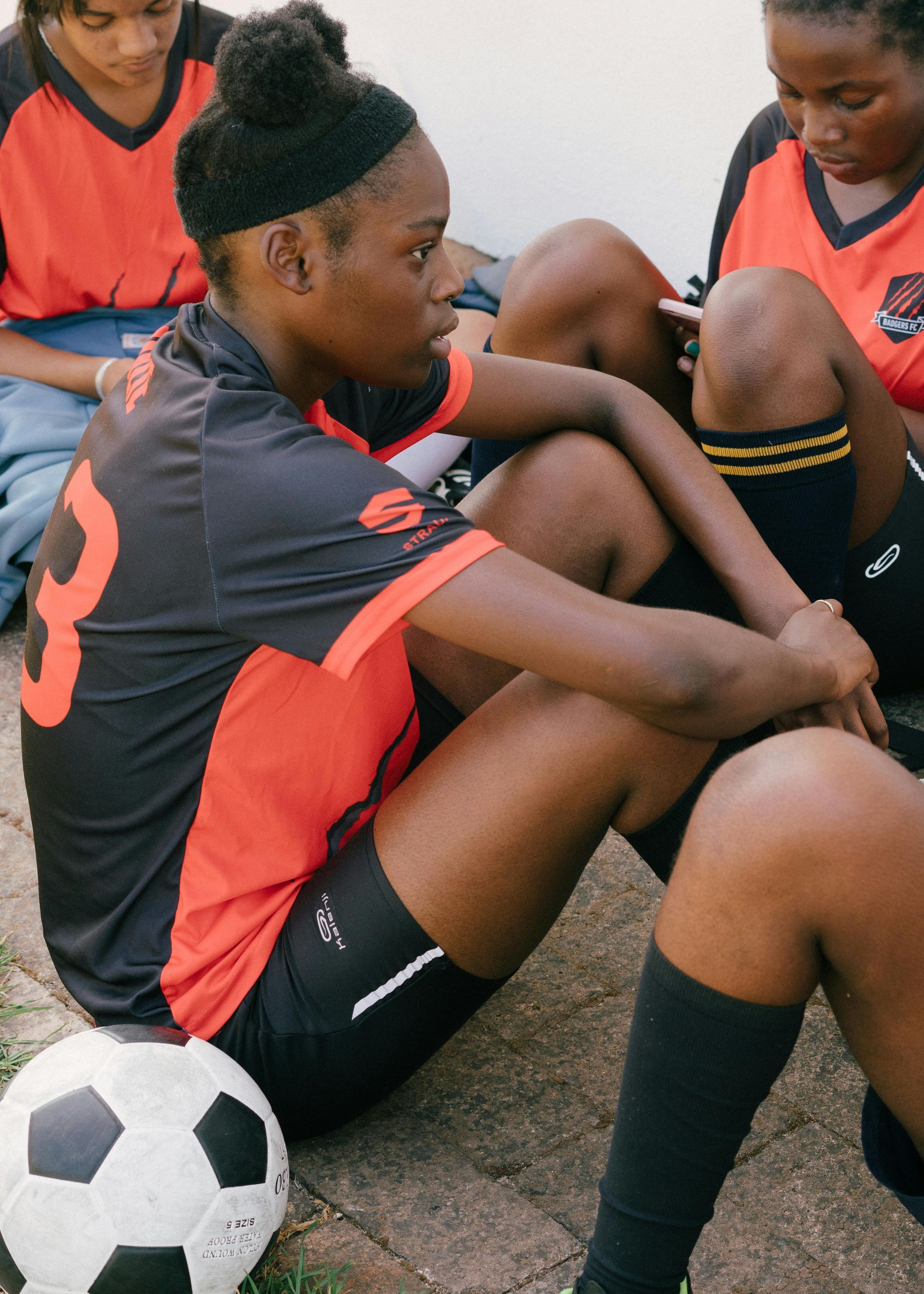Soccer players in black and red uniforms sit on the ground with a soccer ball.
