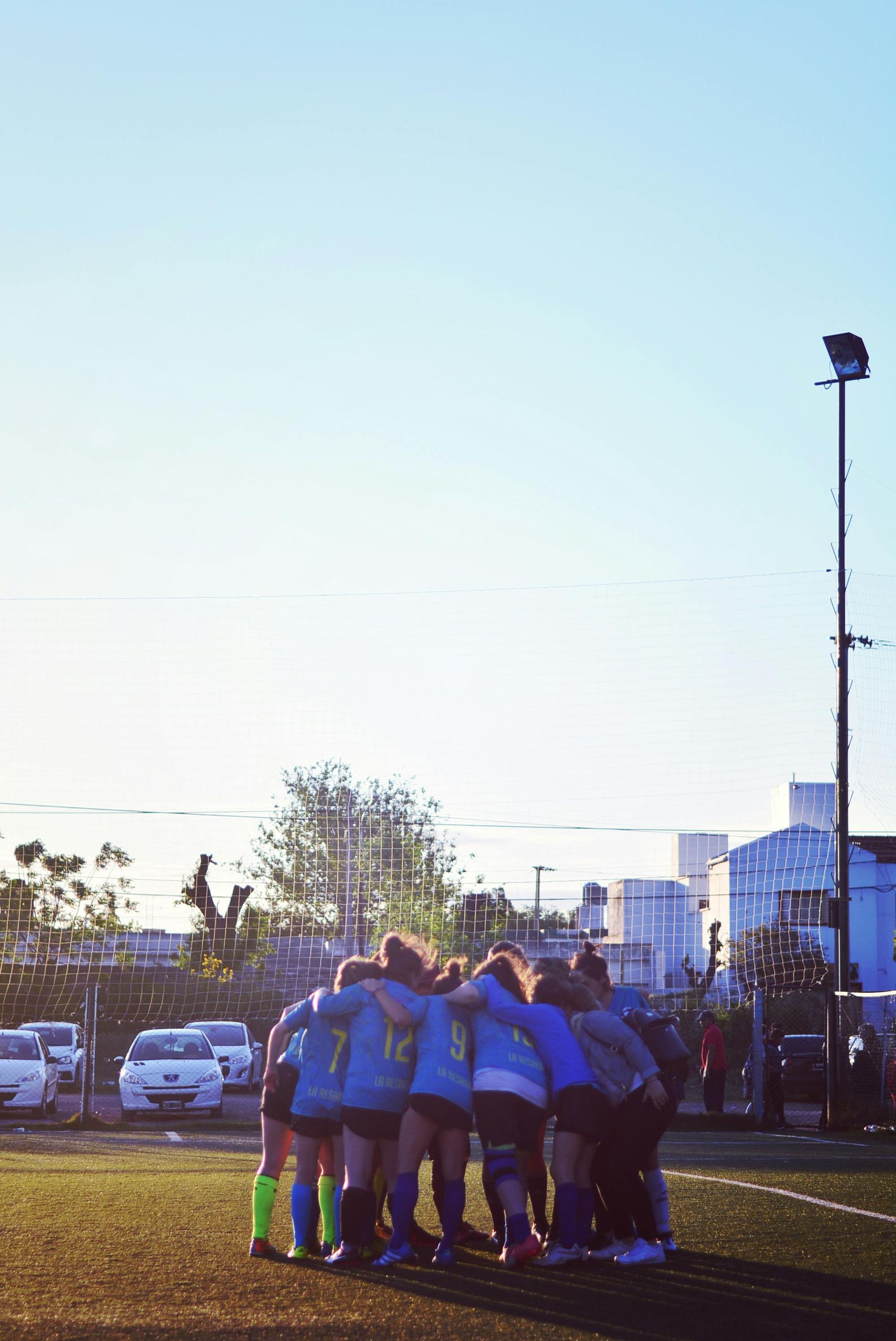 Soccer team huddle on a field, backs turned, light blue uniforms, sunny sky.