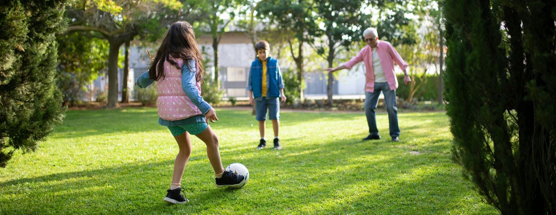 Three people playing soccer on a grassy lawn with trees in the background.