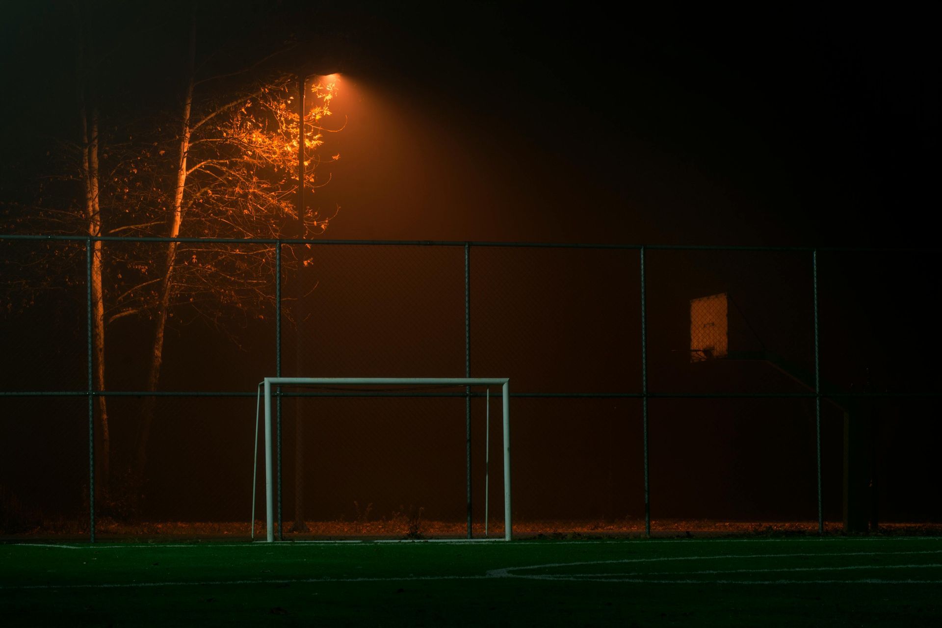 Soccer goal on foggy field under an orange street light at night.