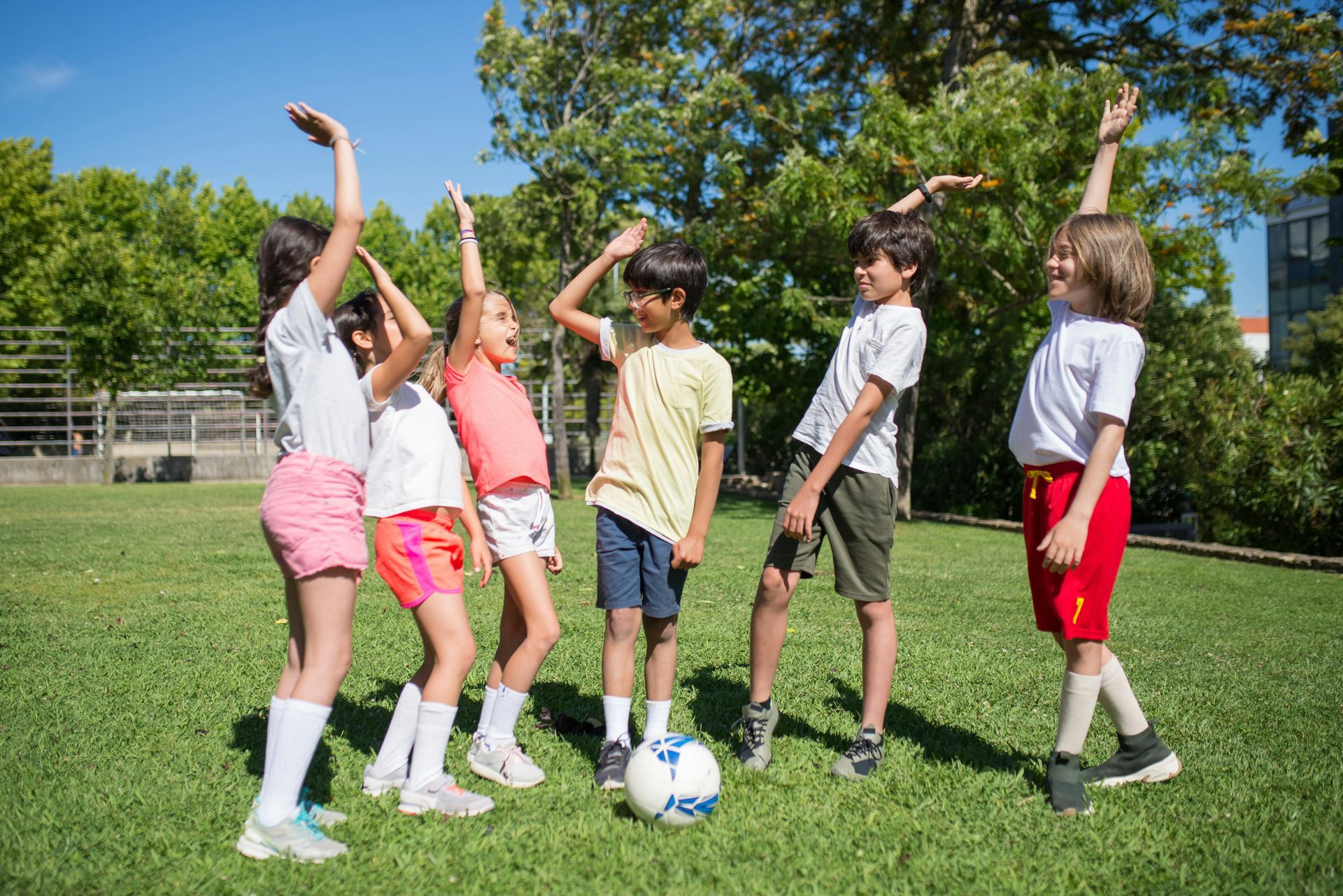 Children on a green lawn with a soccer ball, some raising their arms in play.