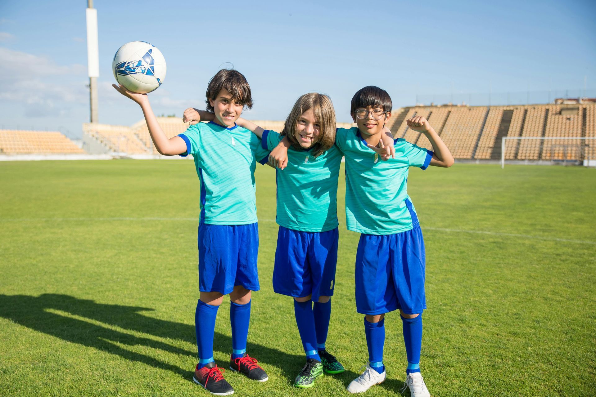 Three soccer players in blue uniforms stand on a green field, one holding the ball, smiling.
