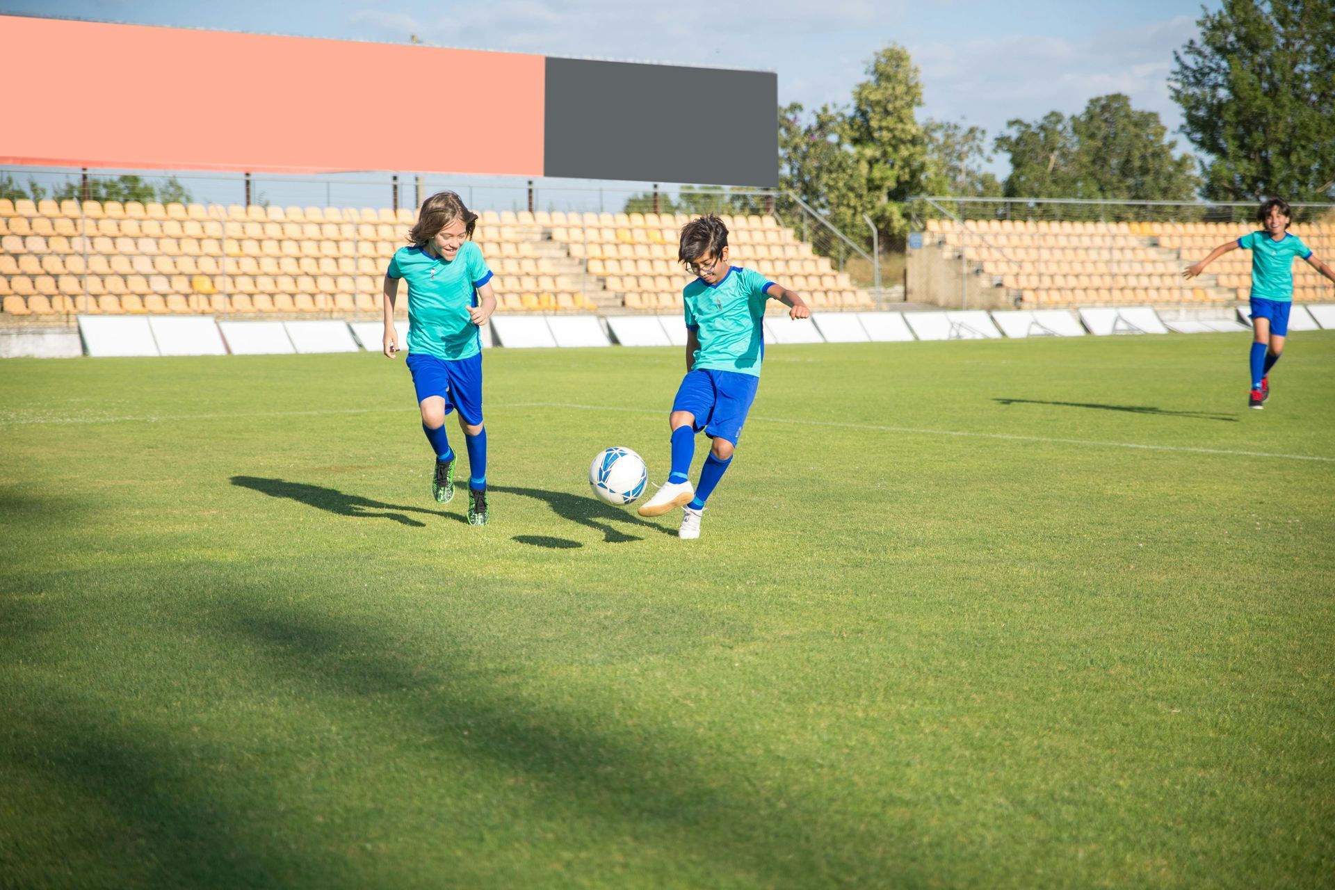 Three soccer players in blue uniforms on a green field. One kicks the ball, the others run.