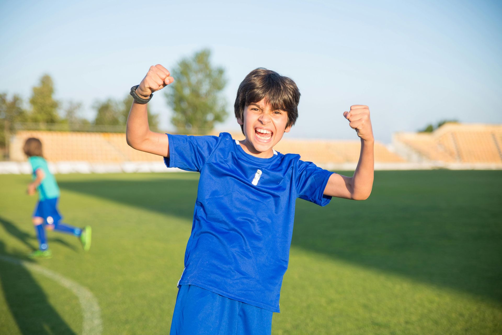 Boy in blue soccer uniform celebrating on field, fists raised, another player in background.