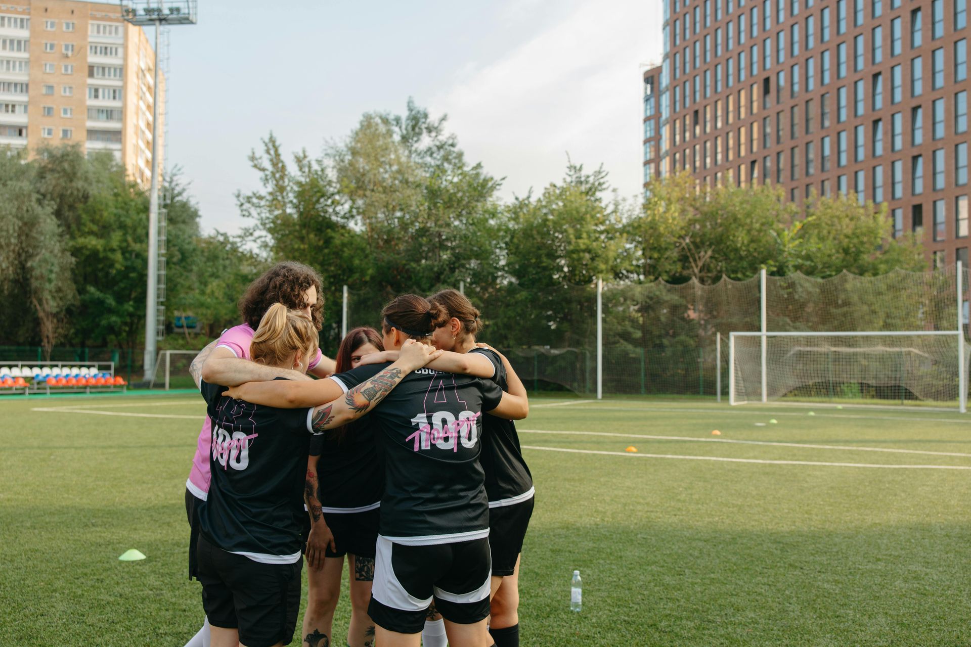 Soccer team huddles together on a green field. Players in black and pink jerseys embrace, building cohesion.