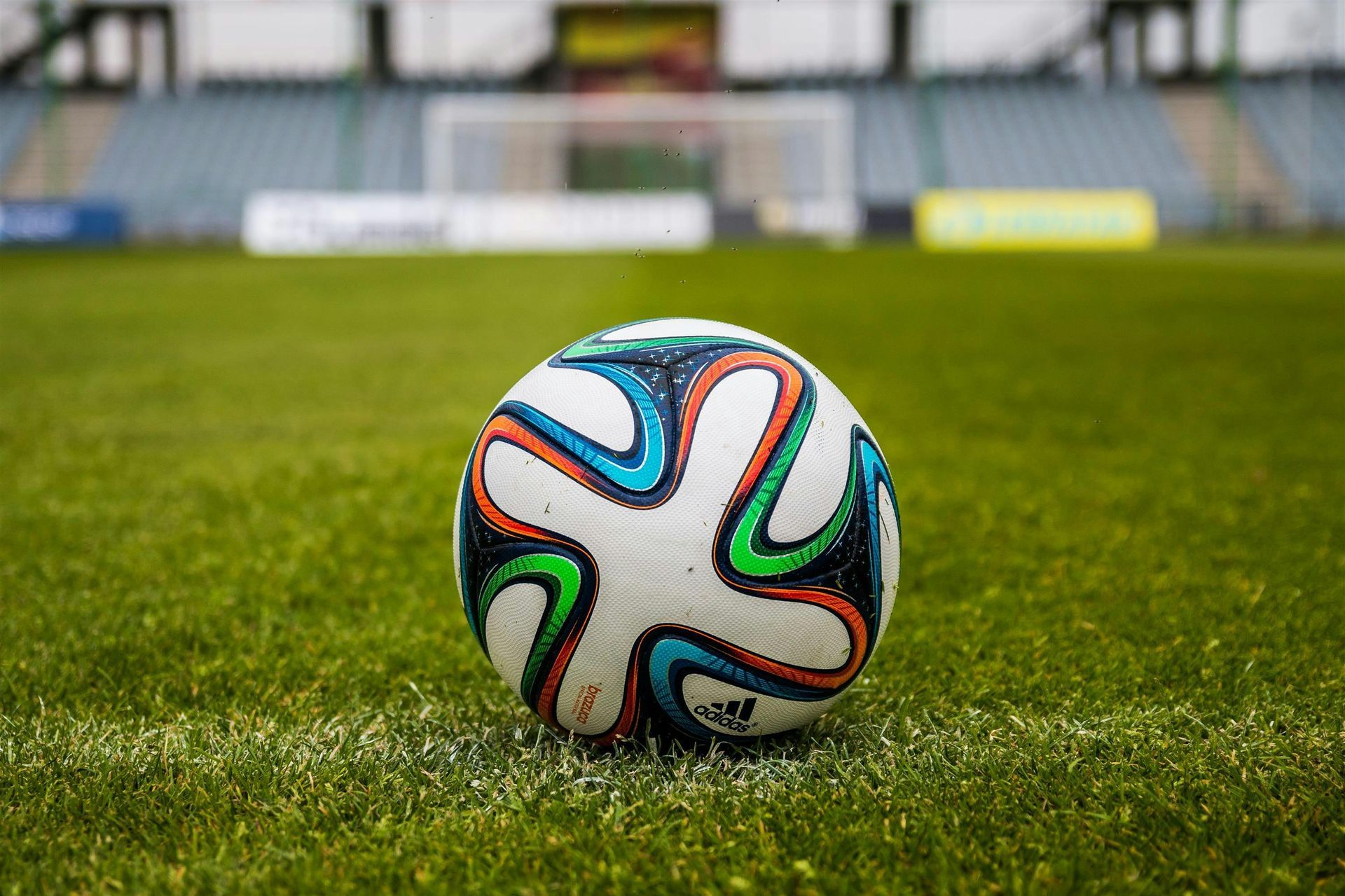 Soccer ball on the field in front of goal. Green grass, stadium seating in the background.