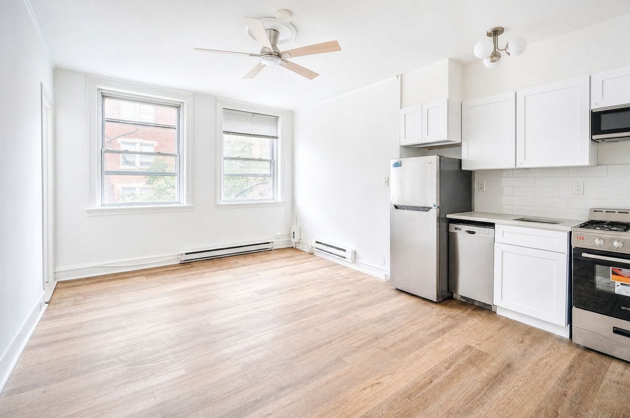Bright empty kitchen-living space with white cabinets, stainless fridge, and large windows showing a wood floor
