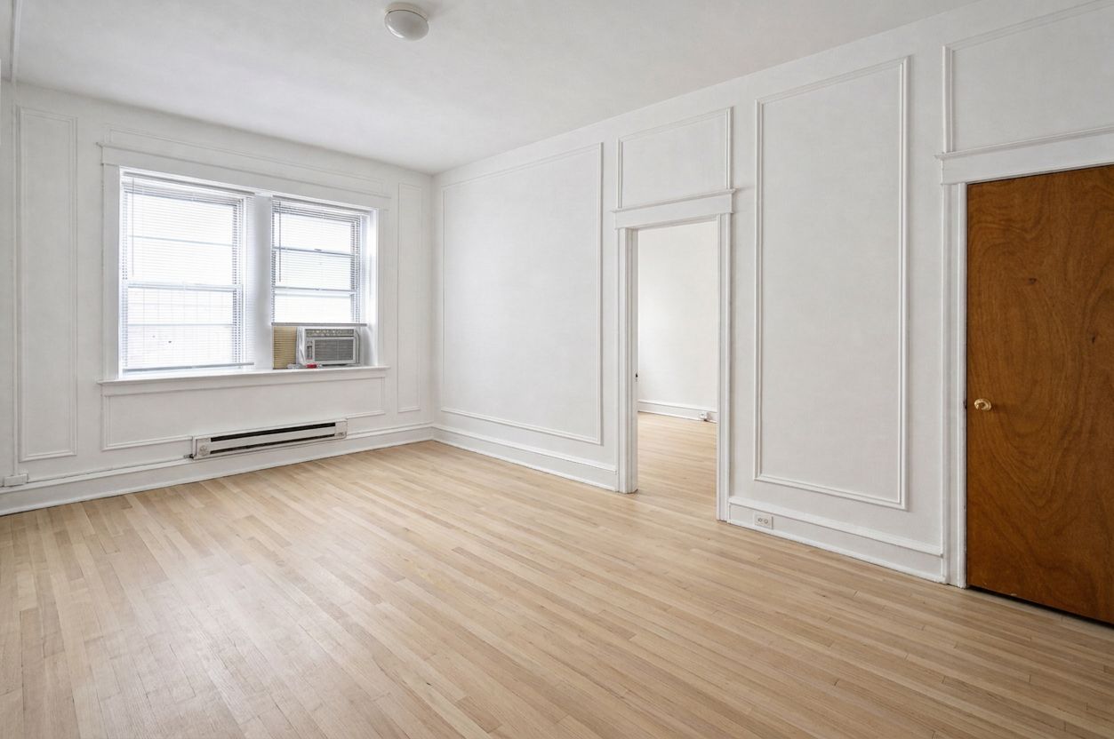 Empty white room with light wood floors, large window, and a wooden door