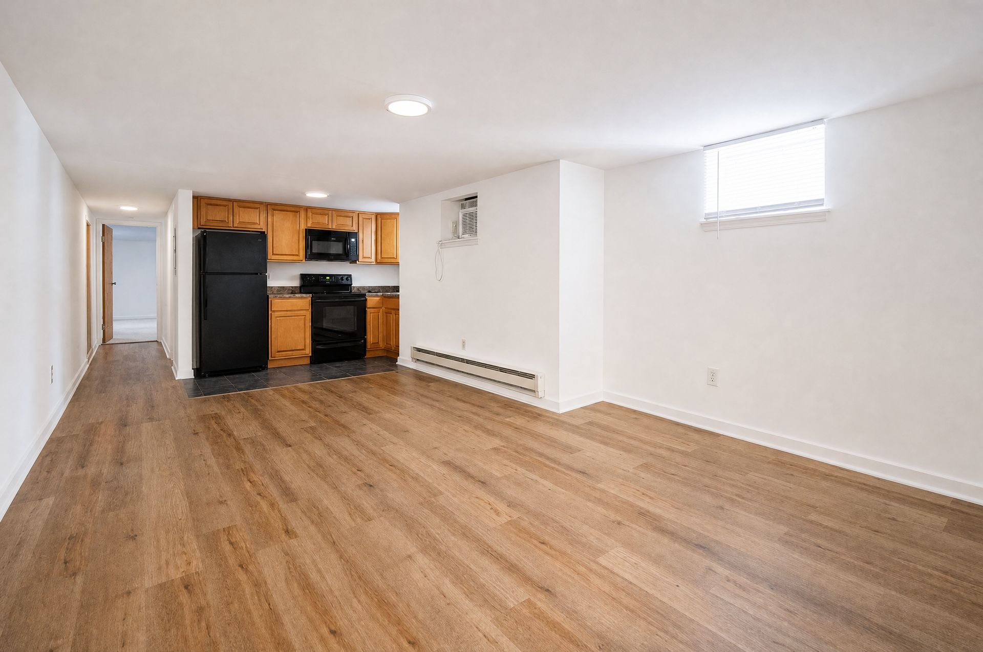 Empty basement living area with wood floors, white walls, and a small kitchen nook.