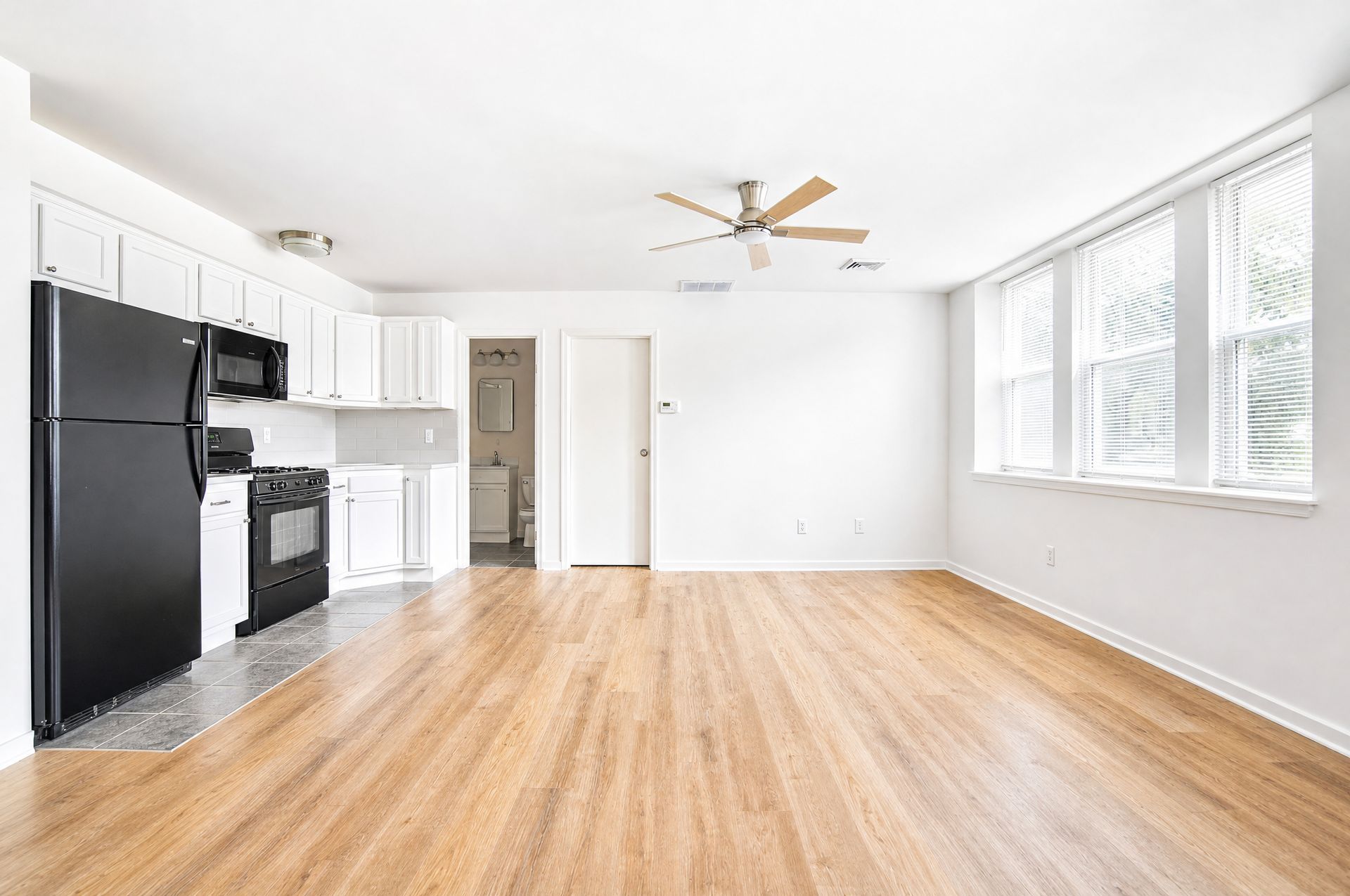 Bright empty kitchen and living area with black appliances, white cabinets, hardwood floors, and large windows.