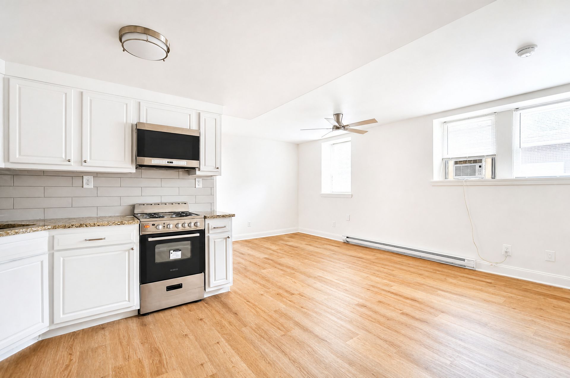 Bright empty kitchen and dining area with white cabinets, stainless stove, hardwood floors, and natural light.