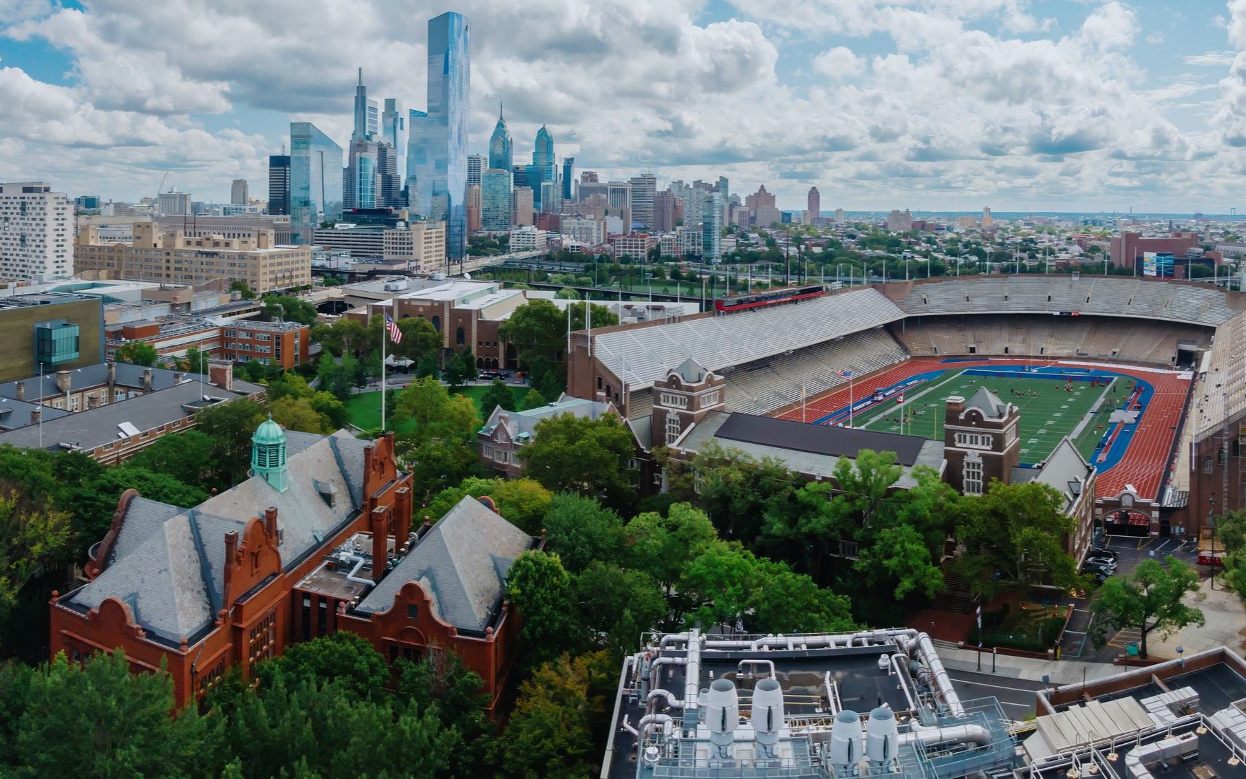 Panoramic city skyline with a red-brick building, stadium, and trees under a cloudy sky