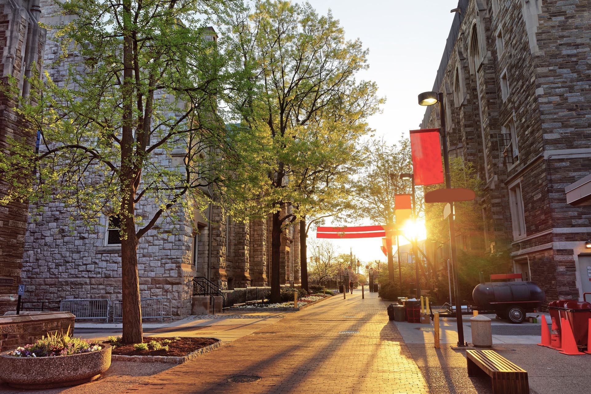 Sunlit stone walkway between buildings with trees and red banners at sunset