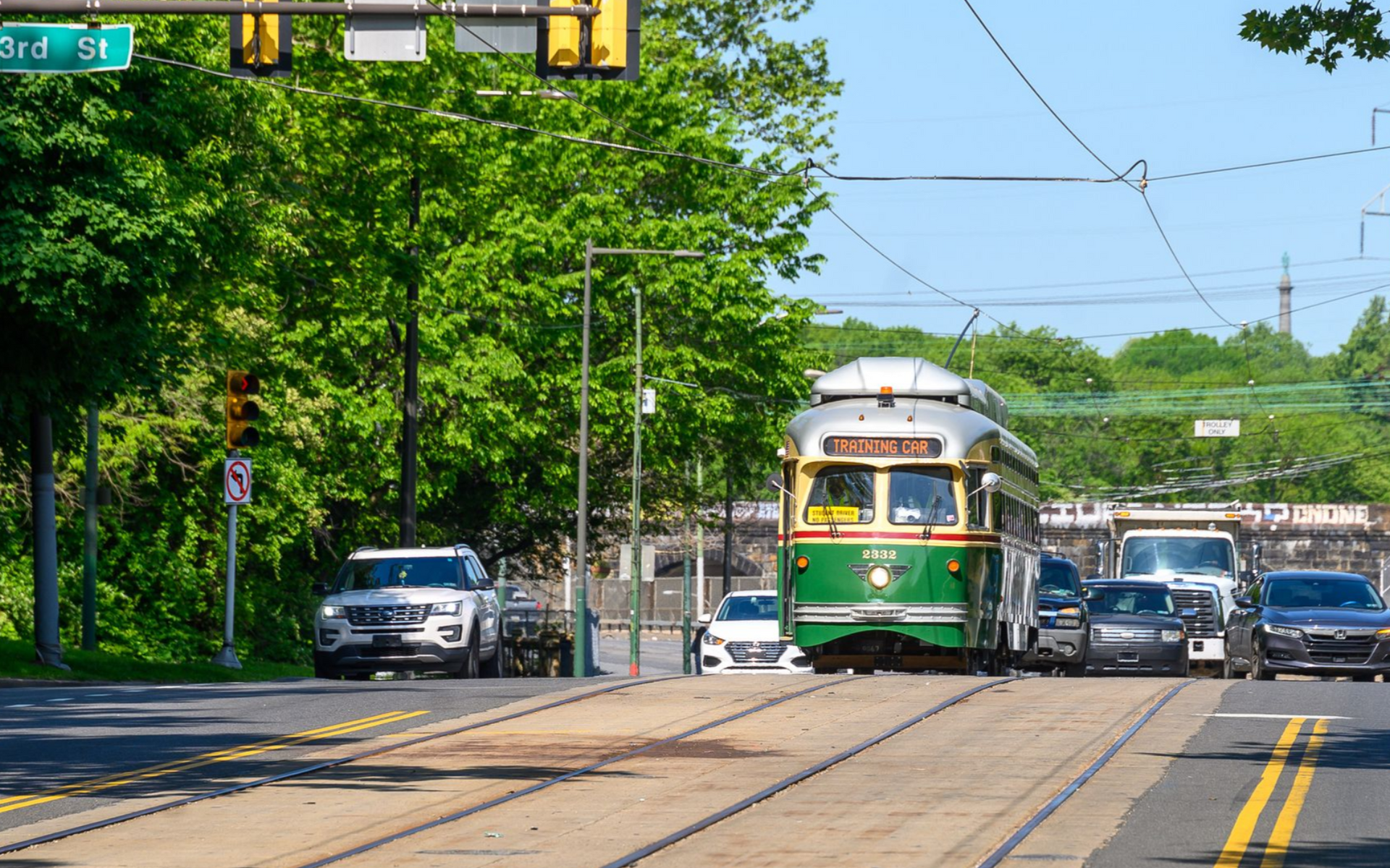 Green-and-white streetcar on tree-lined tracks with cars on a sunny city street