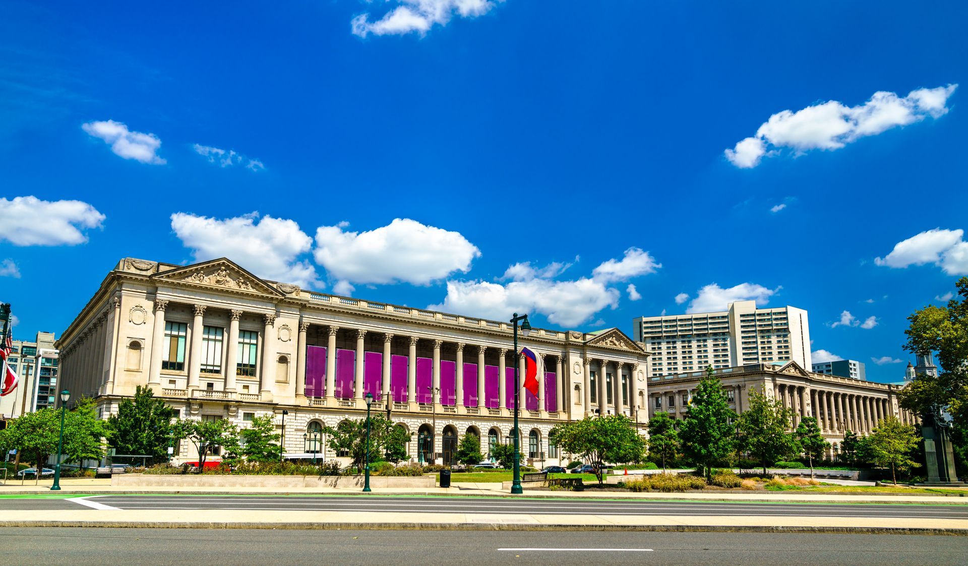 Grand neoclassical building with purple banners under a bright blue sky, viewed from across a wide street