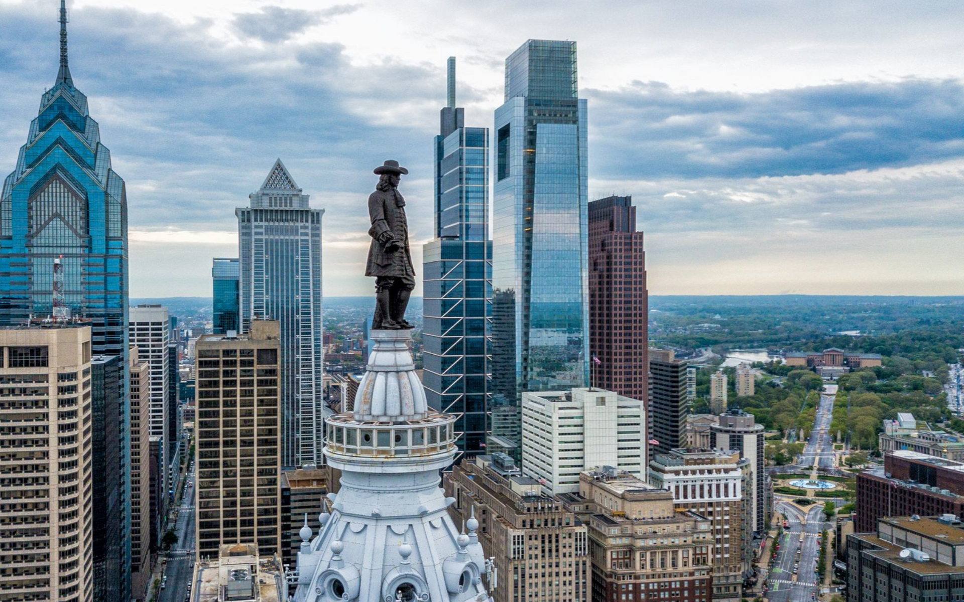 Philadelphia skyline with City Hall statue in foreground and modern skyscrapers under a cloudy blue sky