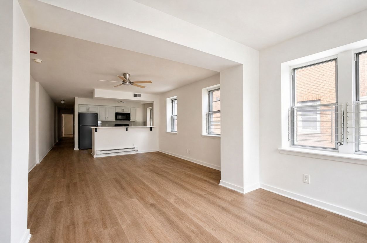 Empty modern apartment living area with wood floors, white walls, and a small kitchen in back
