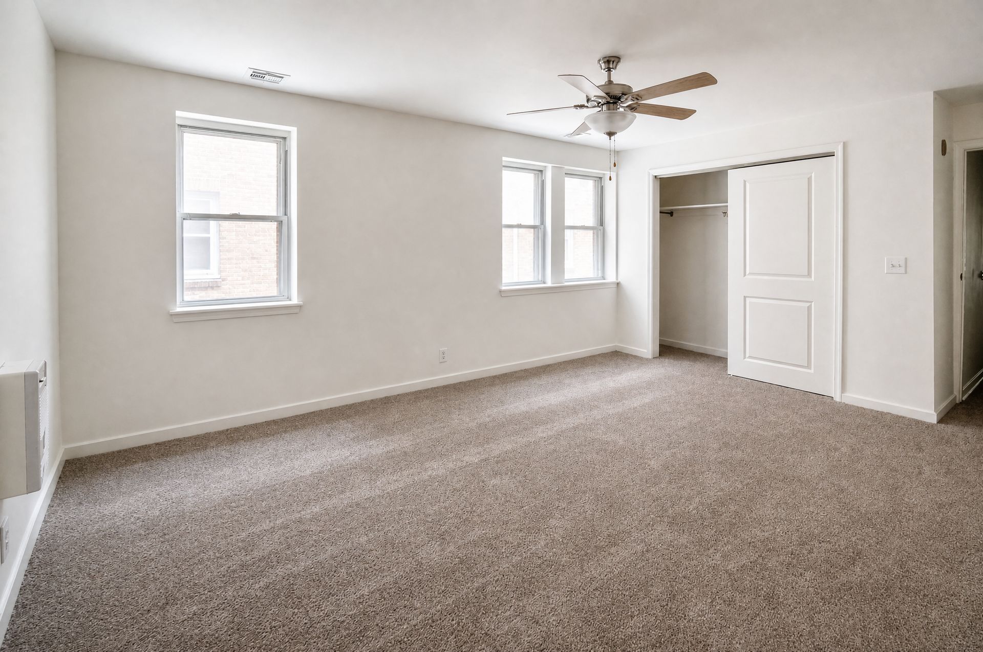 Empty beige carpeted bedroom with white walls, two windows, a ceiling fan, and a closet.