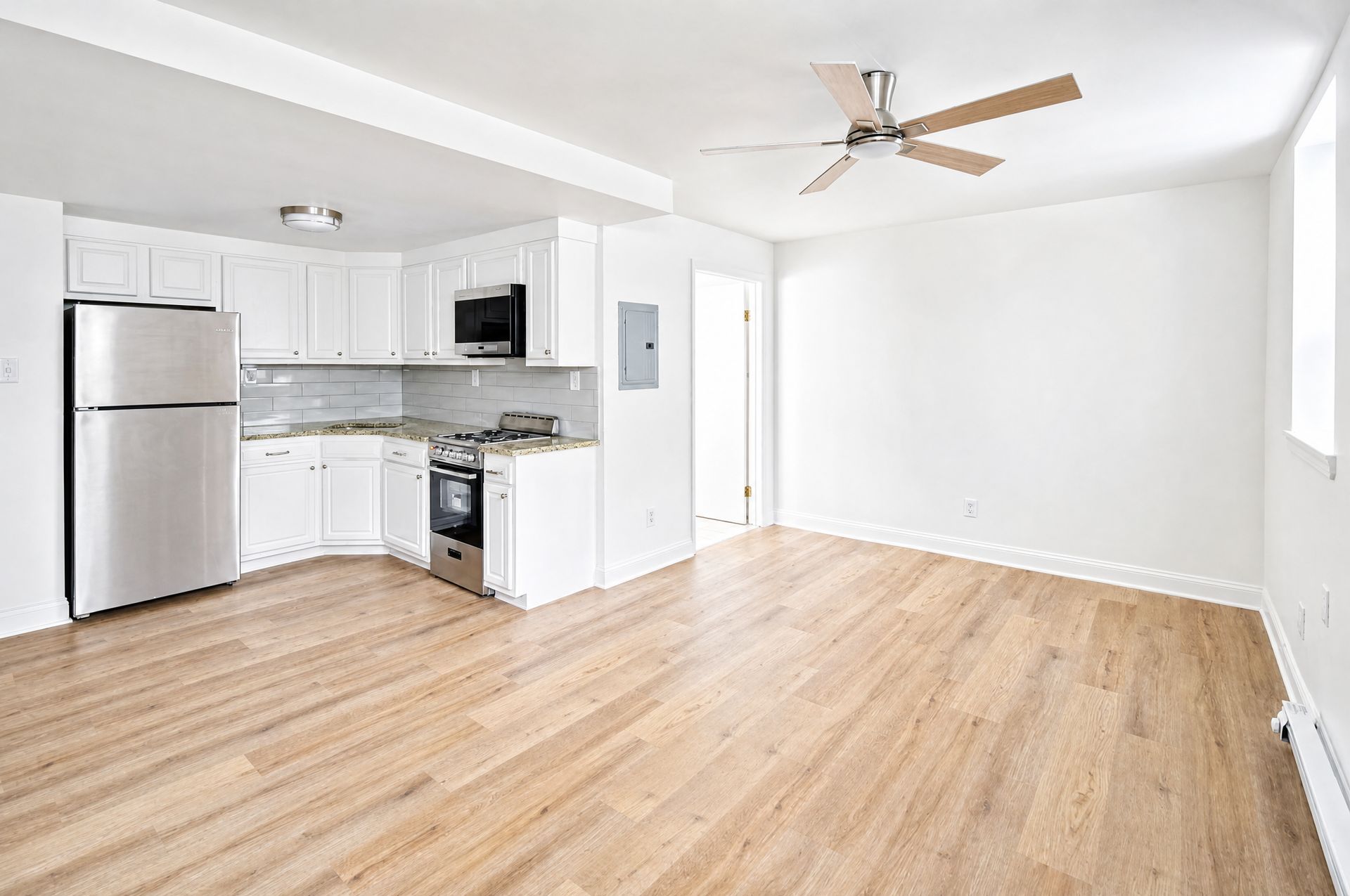 Bright empty living room with light wood floors, white walls, and a small kitchen with stainless fridge.