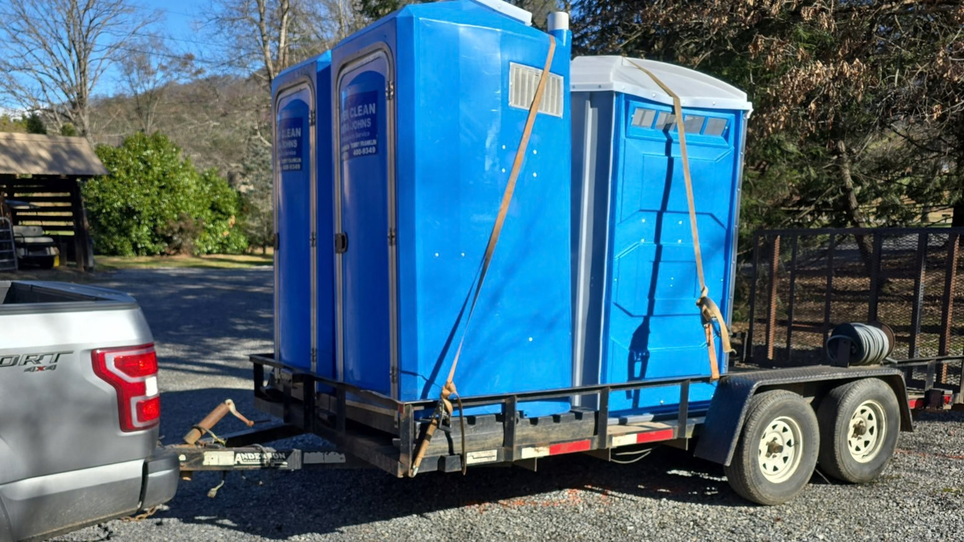 Three blue portable toilets strapped onto a flatbed trailer attached to a pickup truck parked on a gravel lot.