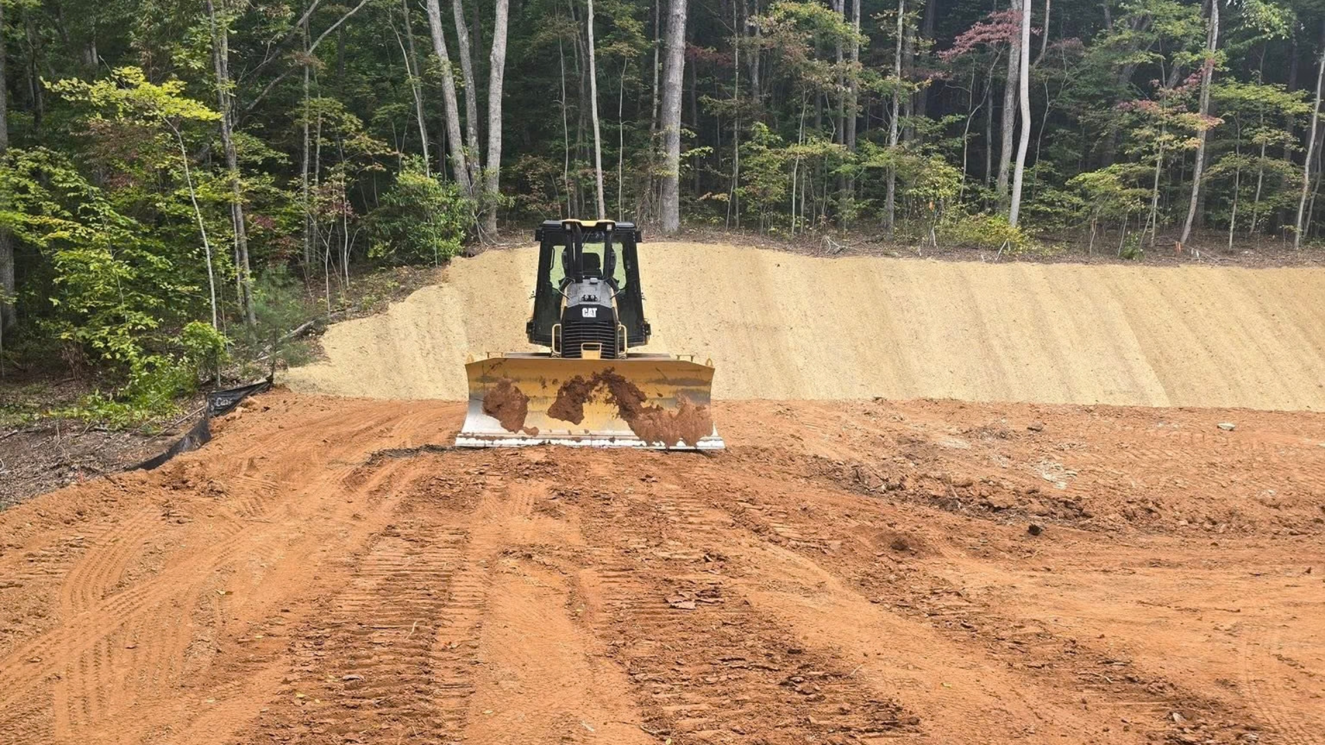 A bulldozer sits on red, cleared earth in front of a graded hillside against a background of trees.