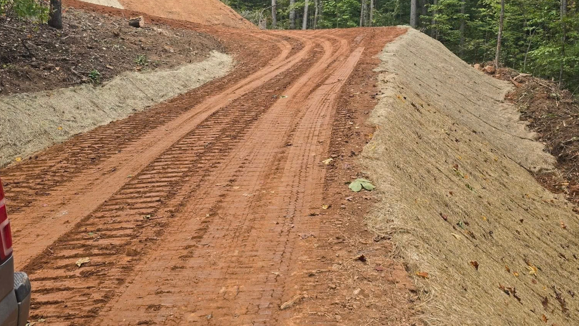 A dirt road winding through a clearing, showing deep tire tracks in the reddish-brown earth and steep, graded banks.
