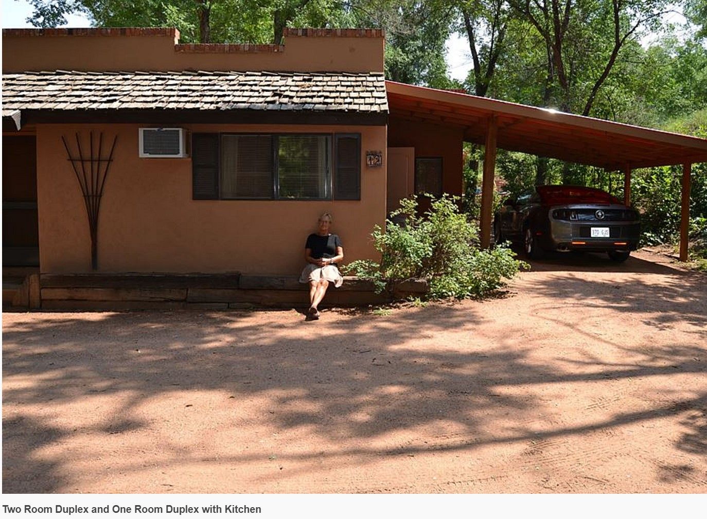 A woman sits in front of a house with a car parked underneath it