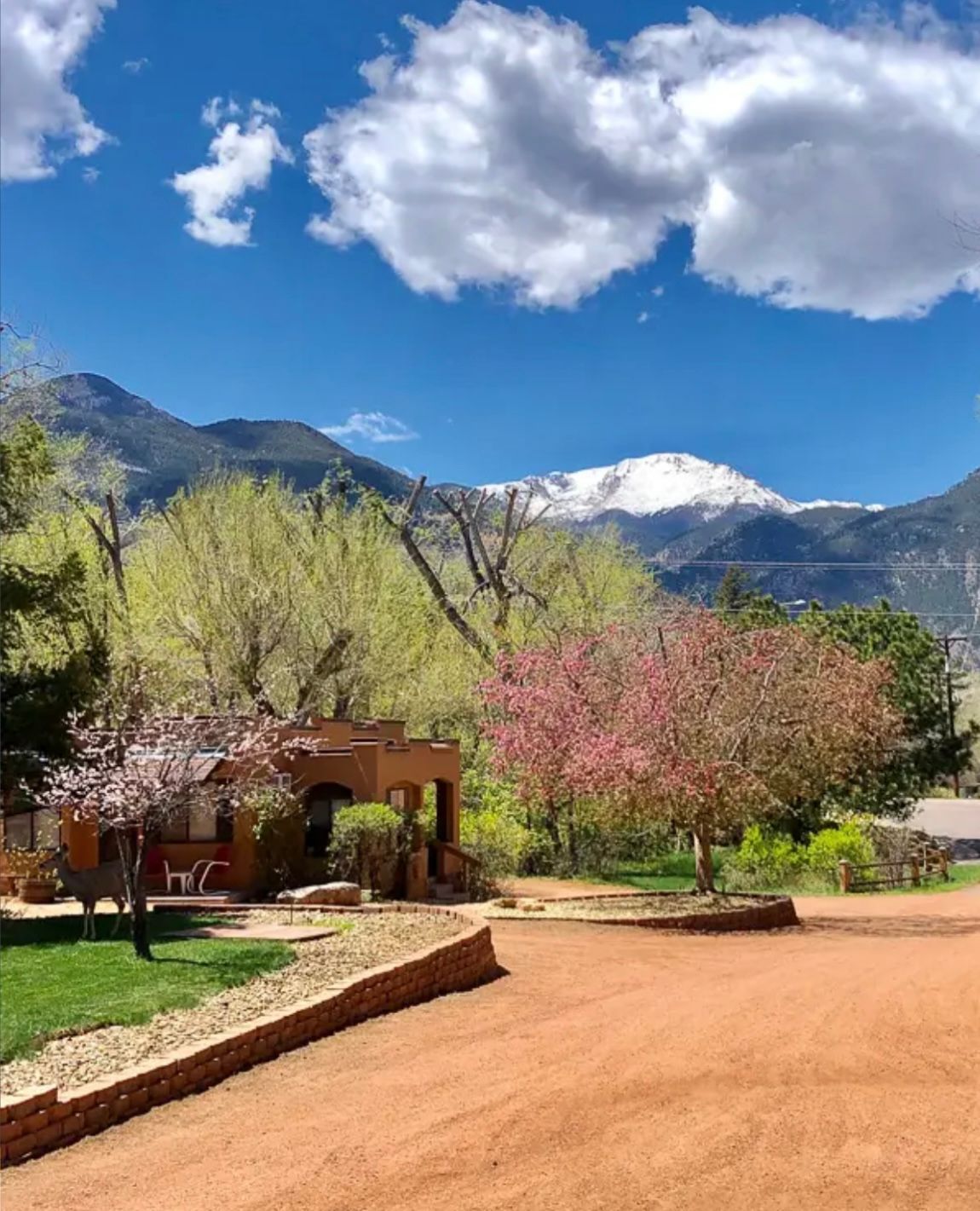 Adobe cottage with flowering trees in front of snow-capped mountains under a blue sky.