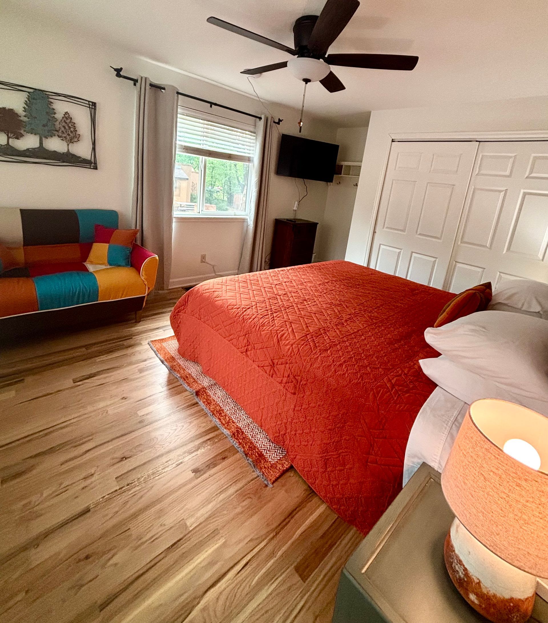 Bedroom with orange bedspread, colorful sofa, wooden floors, and a ceiling fan.