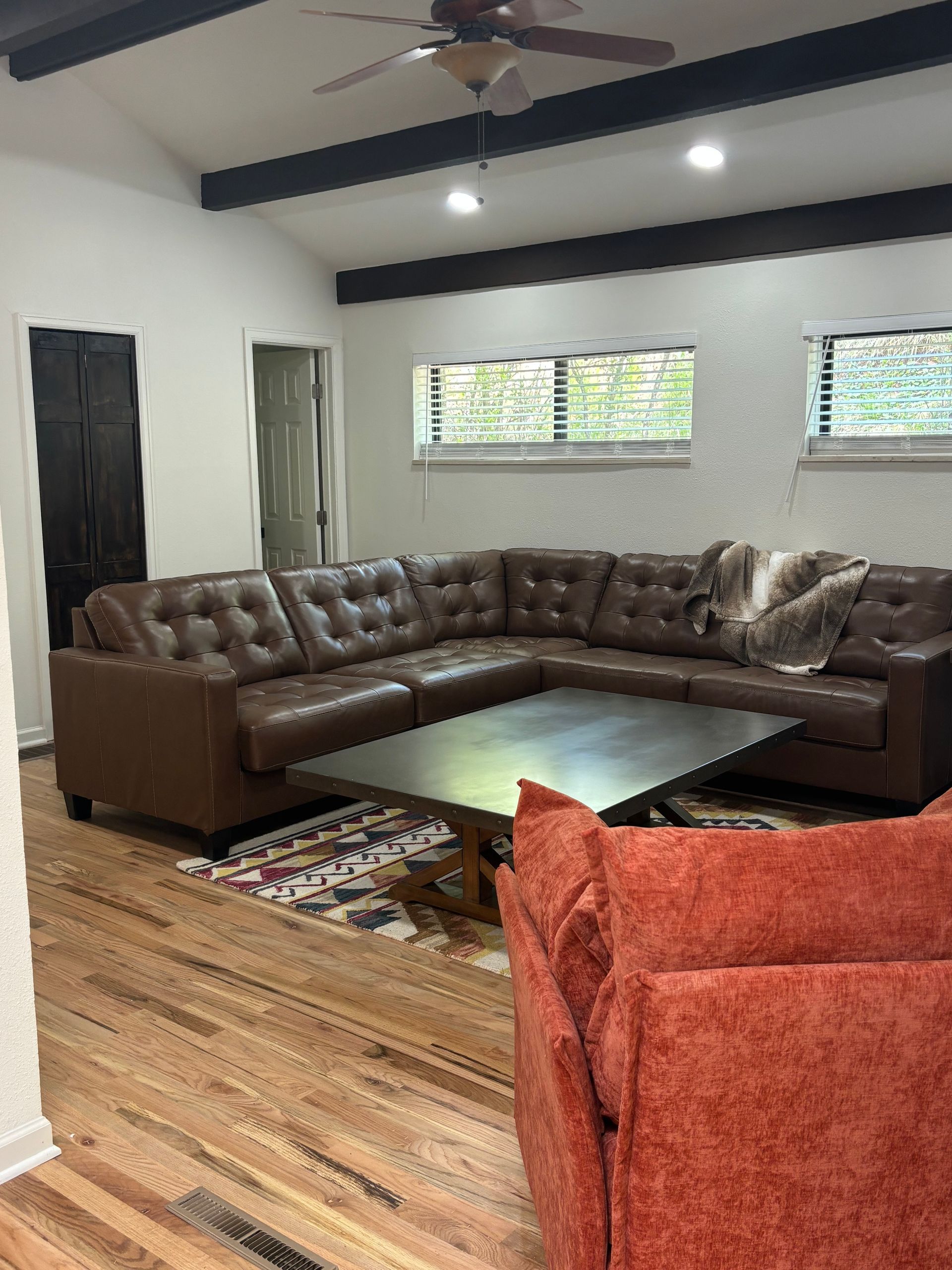 Spacious living room with brown leather sectional, wooden floor, dark ceiling beams, and a red chair.