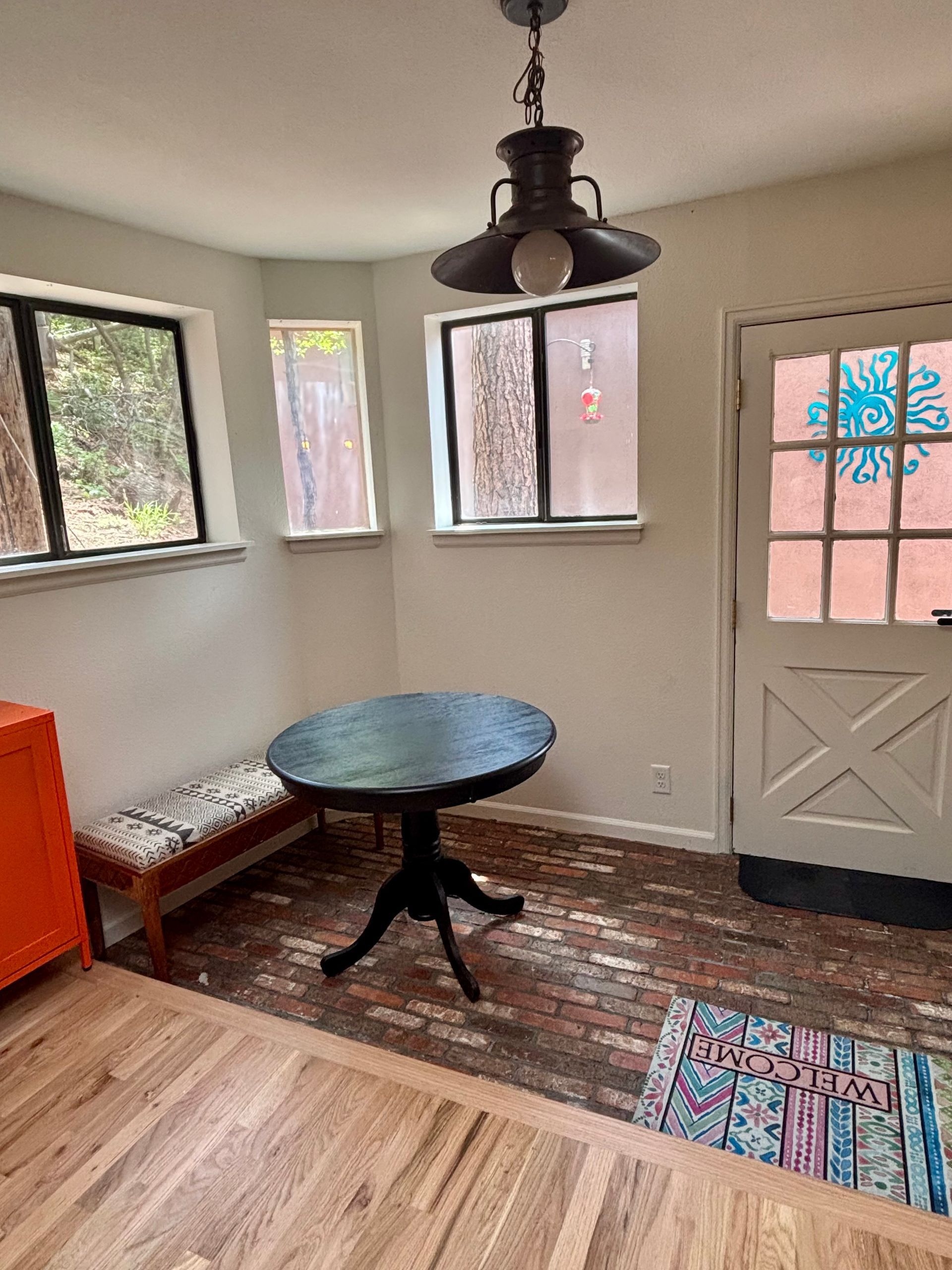 Small dining area with round table, bench, and windows. Brick floor, pendant light.