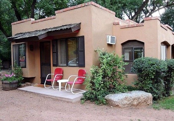 Tan adobe-style cottage with red chairs on porch, air conditioner, and shrubbery.