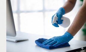 Person in blue gloves cleaning a desk with a spray bottle and microfiber cloth.