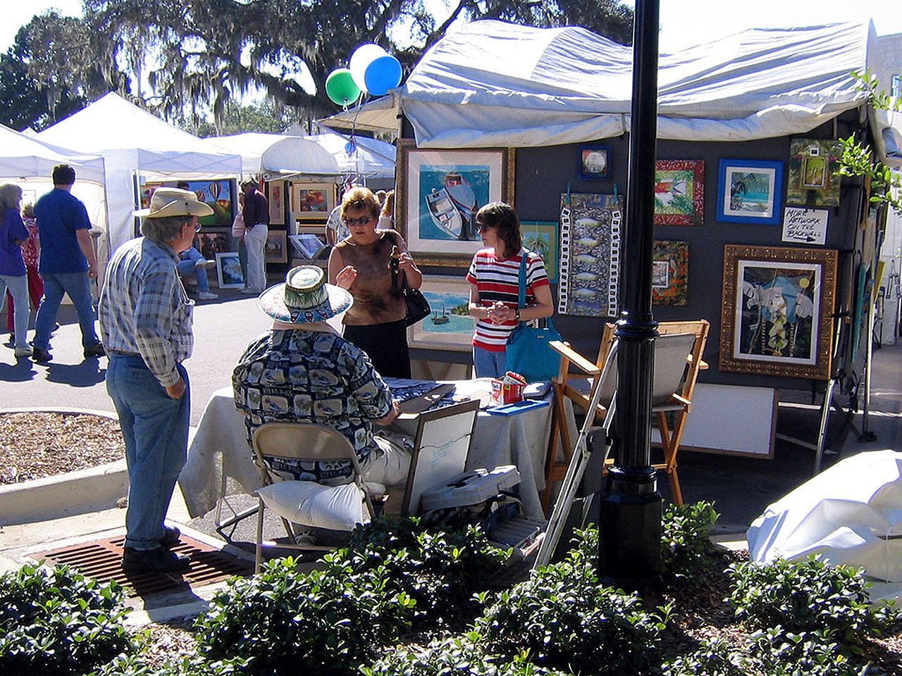 a man in a hat sits at a table in front of a tent with paintings on it