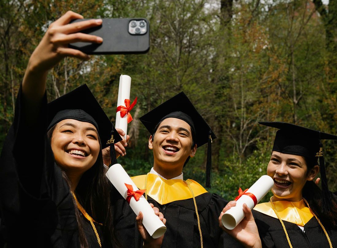 Three graduates in caps and gowns smile for a selfie outdoors, holding diplomas.