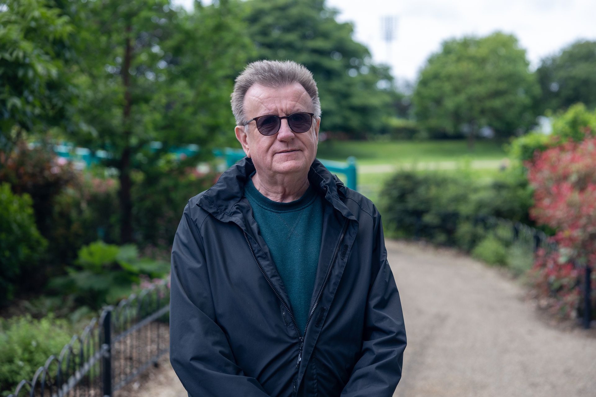 A man wearing sunglasses and a black jacket is standing in a park.