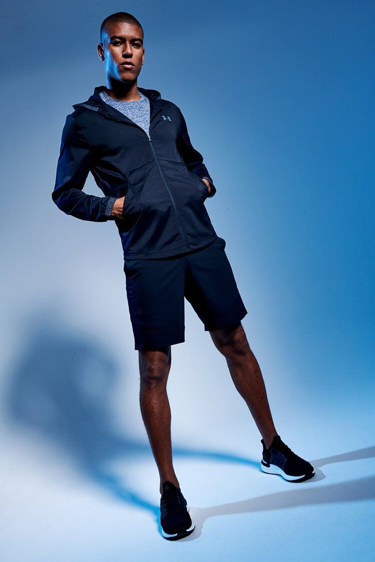 A man in a black jacket and shorts is standing in front of a blue background.