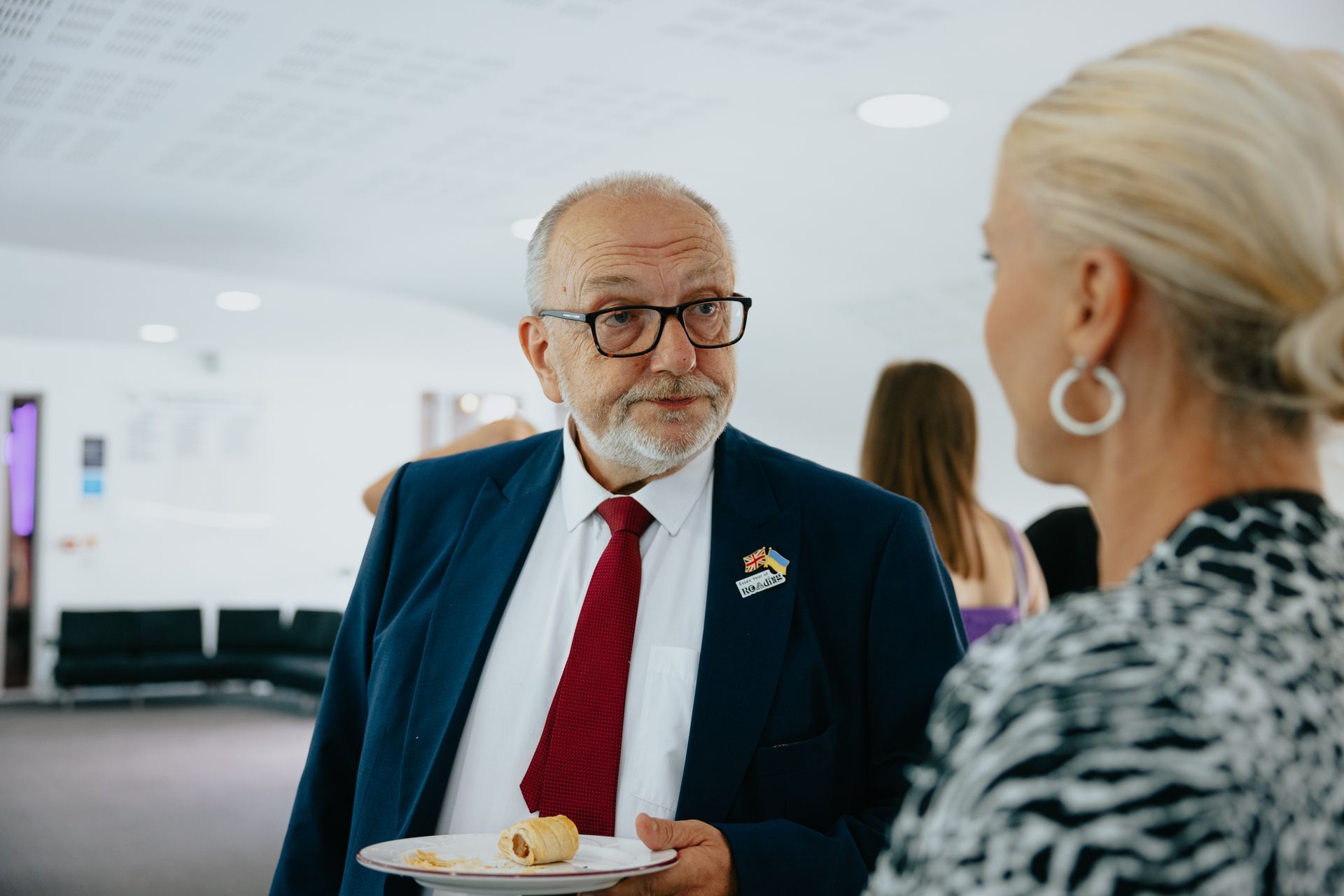 A man in a suit and tie is holding a plate of food and talking to a woman.